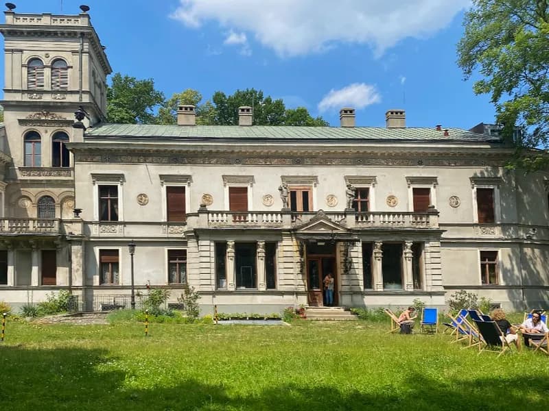 A large, ornate historical building with a prominent tower on the left, an extensive green lawn in front, and several people relaxing in lounge chairs on the right side of the lawn.