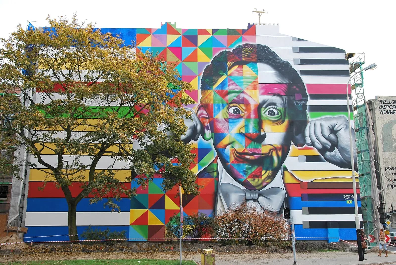 A large colorful mural of a man's face with a shocked expression, wearing a bowtie and divided into geometric patterns on one side and black and white on the other, covering the side of a building next to a tree.